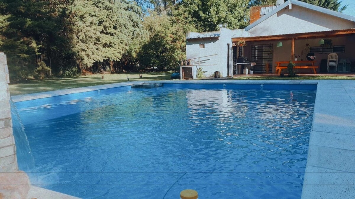 A serene swimming pool is framed by lush greenery, providing a peaceful retreat. The pool's clear water reflects the surrounding trees and features a shallow area with a gentle waterfall. A well-maintained patio area lies adjacent to the pool, enhancing the outdoor space.