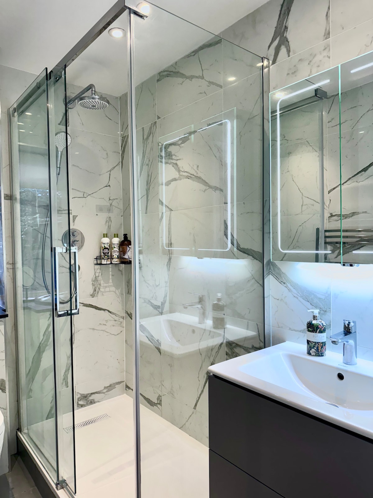 The modern bathroom is characterized by elegant marble walls and a glass shower enclosure. A mirror with built-in lighting offers a bright reflection, while a sleek vanity provides storage space. Amenities include a towel rack, soap dispensers, and a modern faucet.