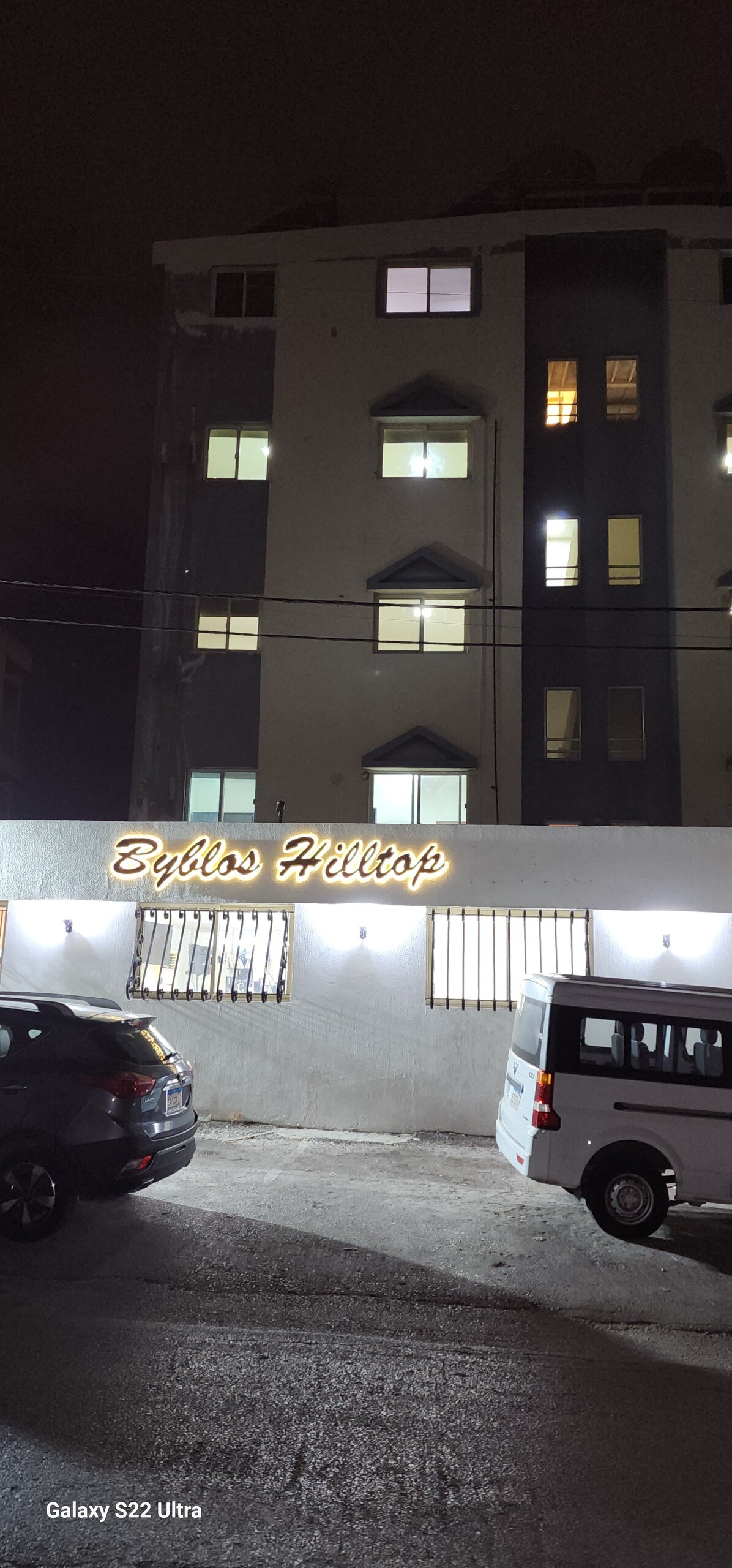 The exterior of the building is captured at night, showcasing multiple illuminated windows and a warm sign that reads 'By Bliss Hilltop.' Vehicles are parked in front, and the structure features a mix of modern and traditional architectural elements.