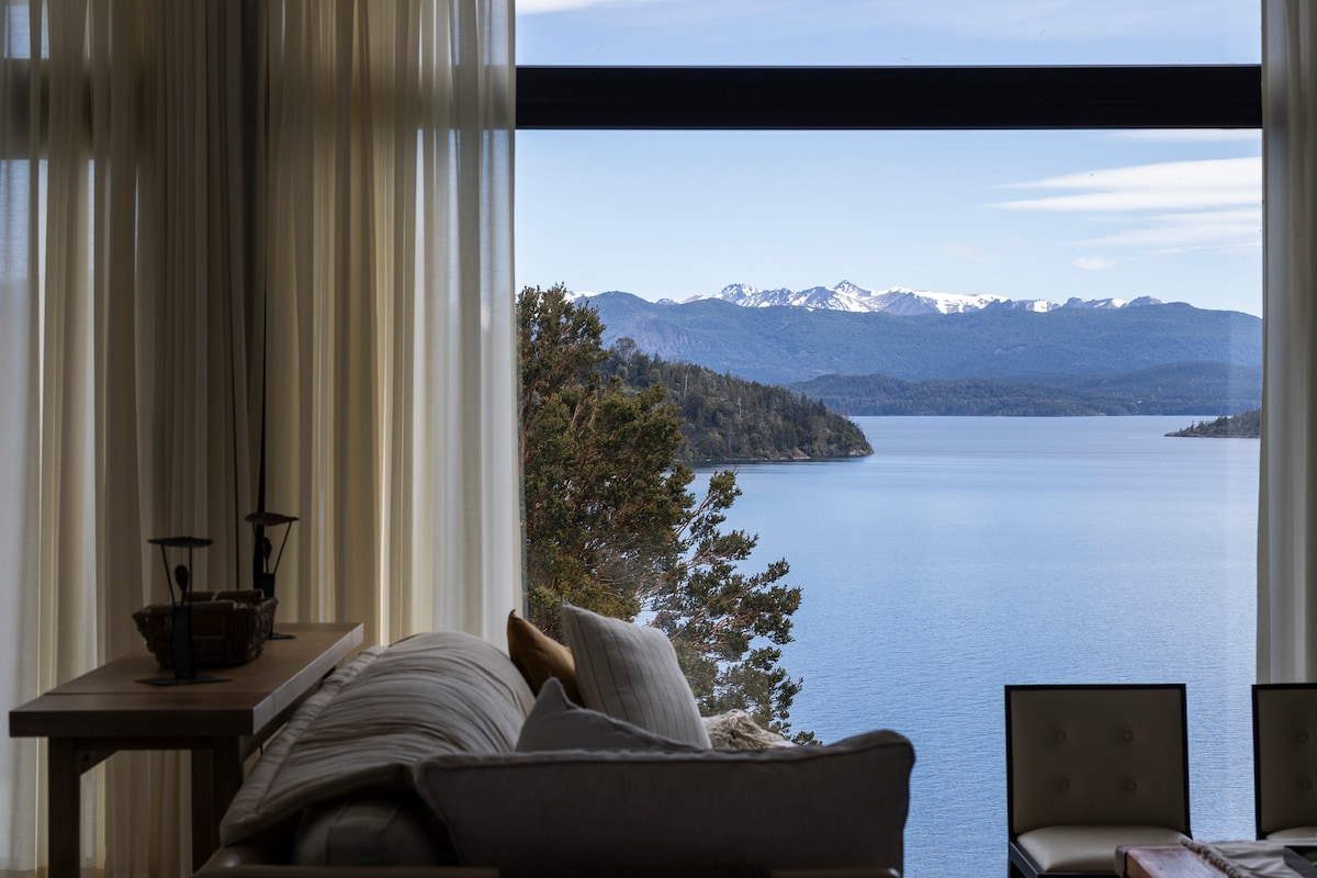 A spacious terrace of 60 square meters features an exclusive hot tub and barbecue area. Clear views of the Nahuel Huapi Lake and mountains are visible, with outdoor furniture arranged for relaxation. This inviting space offers an ideal spot for enjoying the surrounding natural beauty.