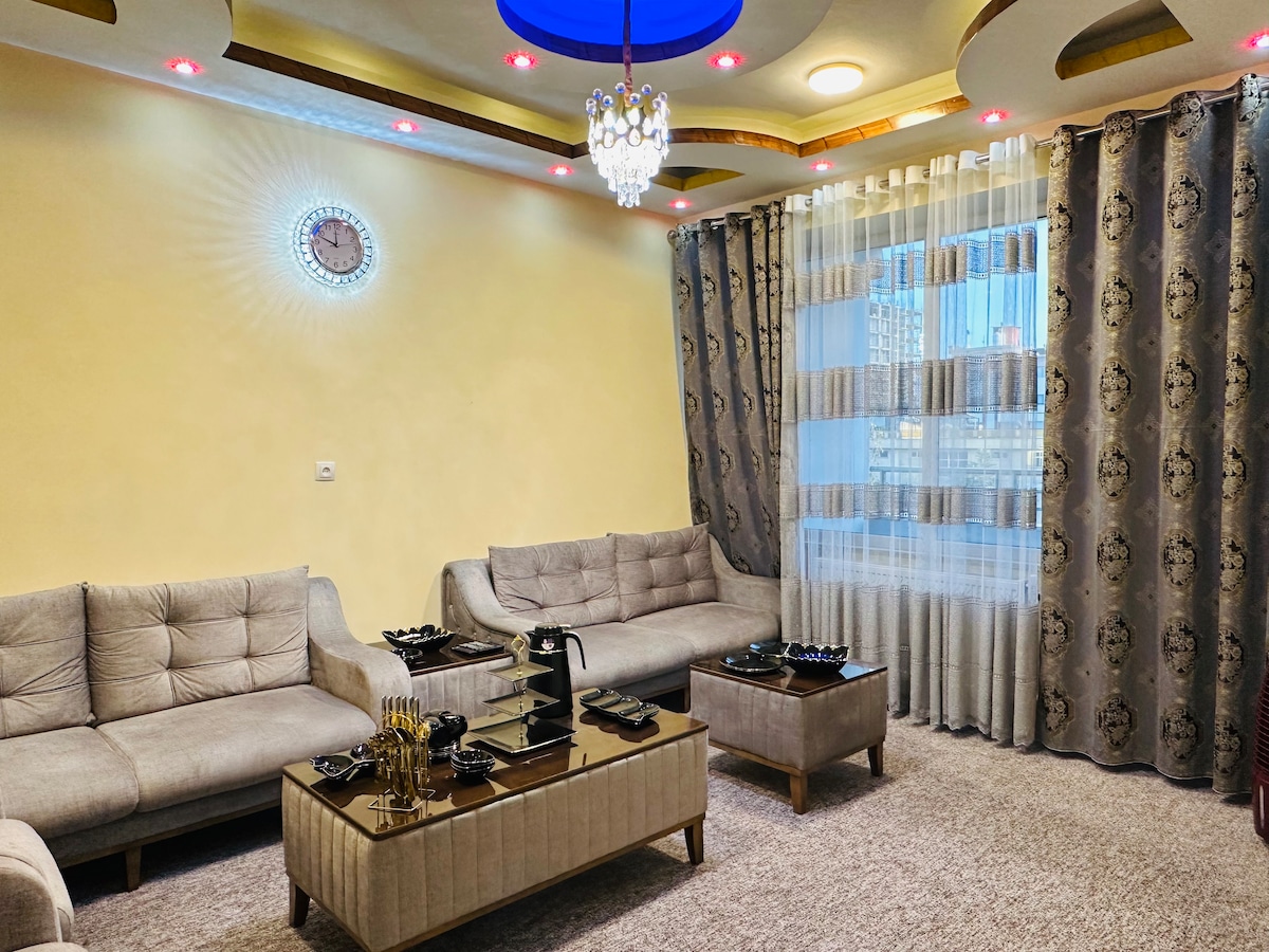 A well-appointed living room features two gray upholstered sofas and a central coffee table. Elegant window treatments frame a large window, allowing natural light to fill the space. Decorative lighting enhances the room, and a clock is mounted on the wall, adding charm.