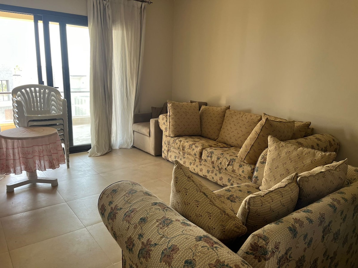 A light-filled living area showcases a floral-patterned sofa and armchair, offering comfortable seating. A small round table is placed nearby, and sheer curtains gently frame the glass doors leading to a balcony, enhancing the space's openness.