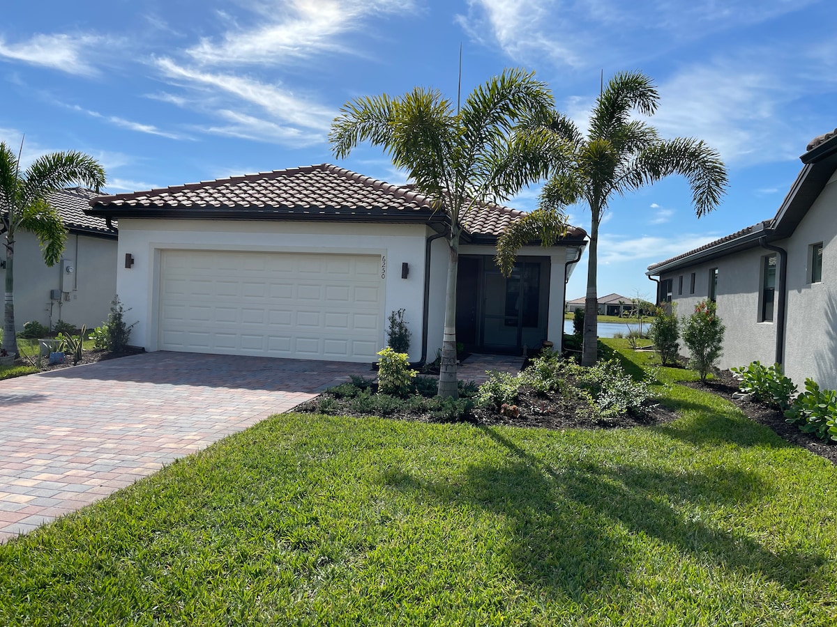 A single-story home is shown with a tiled driveway leading to a two-car garage. Lush green landscaping surrounds the house, including well-maintained shrubs and palm trees under a clear blue sky.