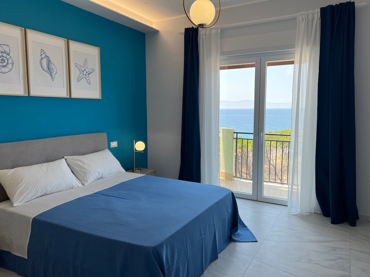 A modern bedroom features a spacious bed with crisp white linens, complemented by a deep blue wall and coastal-themed artwork. Large sliding doors offer a clear view of the sea and access to a private balcony, while soft curtains frame the natural light.