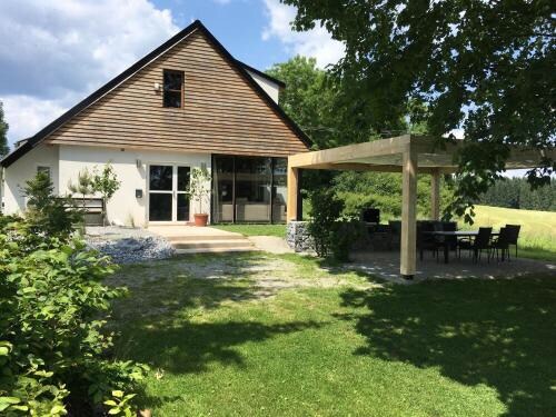 A modern house with a sloped roof is surrounded by lush greenery and a stone pathway. An outdoor seating area is sheltered by a wooden pergola, providing a space for relaxation amid nature. Large windows invite ample natural light into the home.