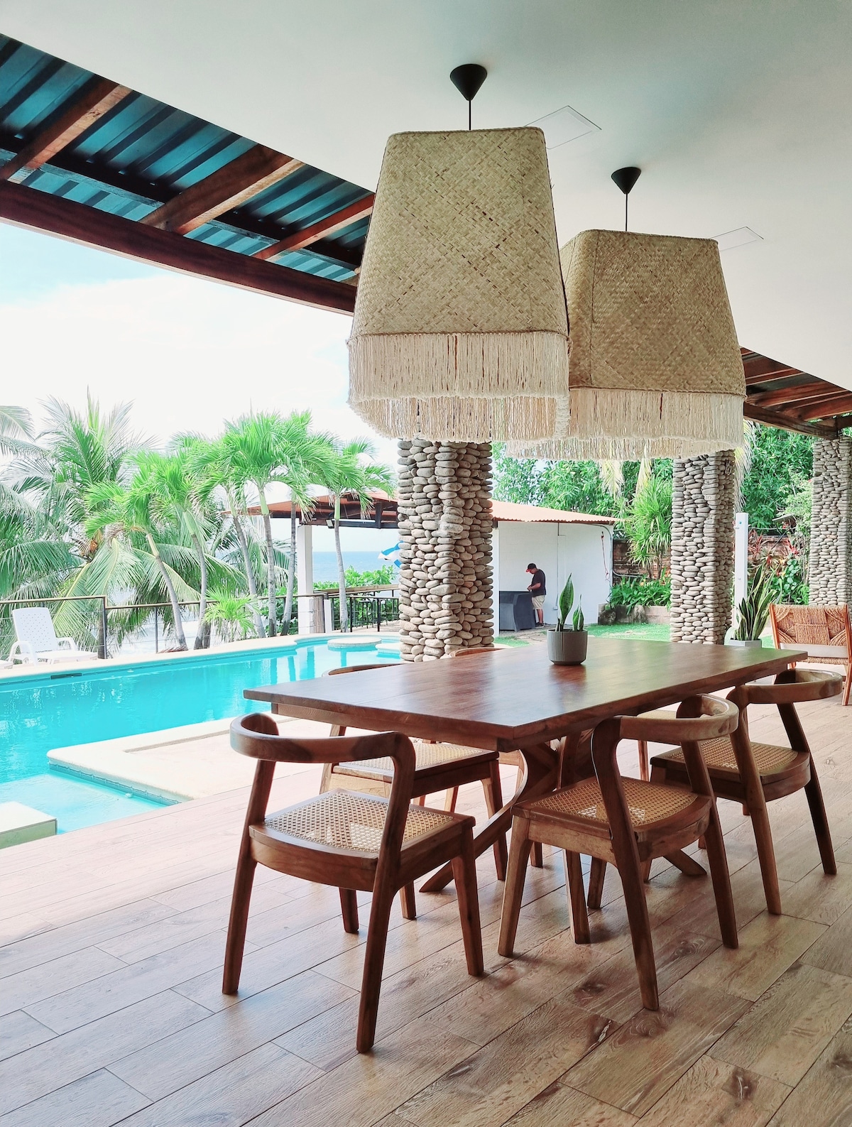 A spacious dining area is situated under woven pendant lights, featuring a rustic wooden table surrounded by four chairs. The backdrop reveals a private swimming pool, lounging area, and lush greenery, enhancing the connection between indoor and outdoor spaces.