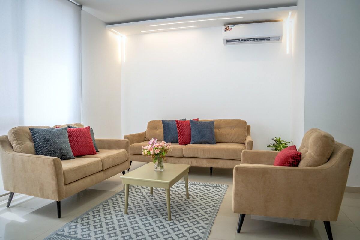 A bright living area is presented with two beige sofas and a matching armchair, all decorated with colorful cushions. A small coffee table is centered on a patterned rug. Natural light enters through a large window, complemented by soft, neutral-colored walls.