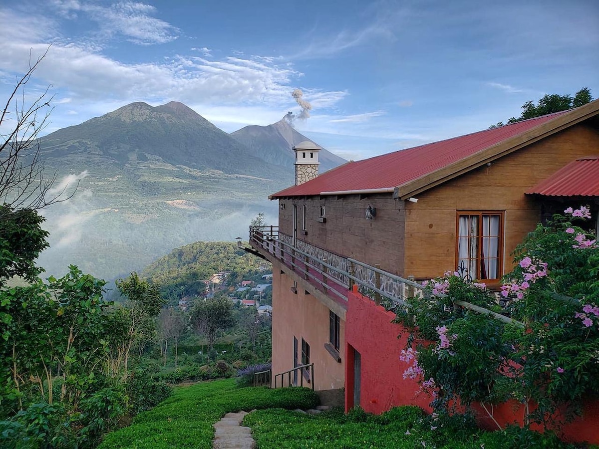 A hillside view of a wooden house features a red roof and large windows. Lush greenery surrounds the property, with blooming flowers adding color. Two prominent volcanoes rise in the background, partially shrouded in mist, suggesting a serene atmosphere in a natural setting.