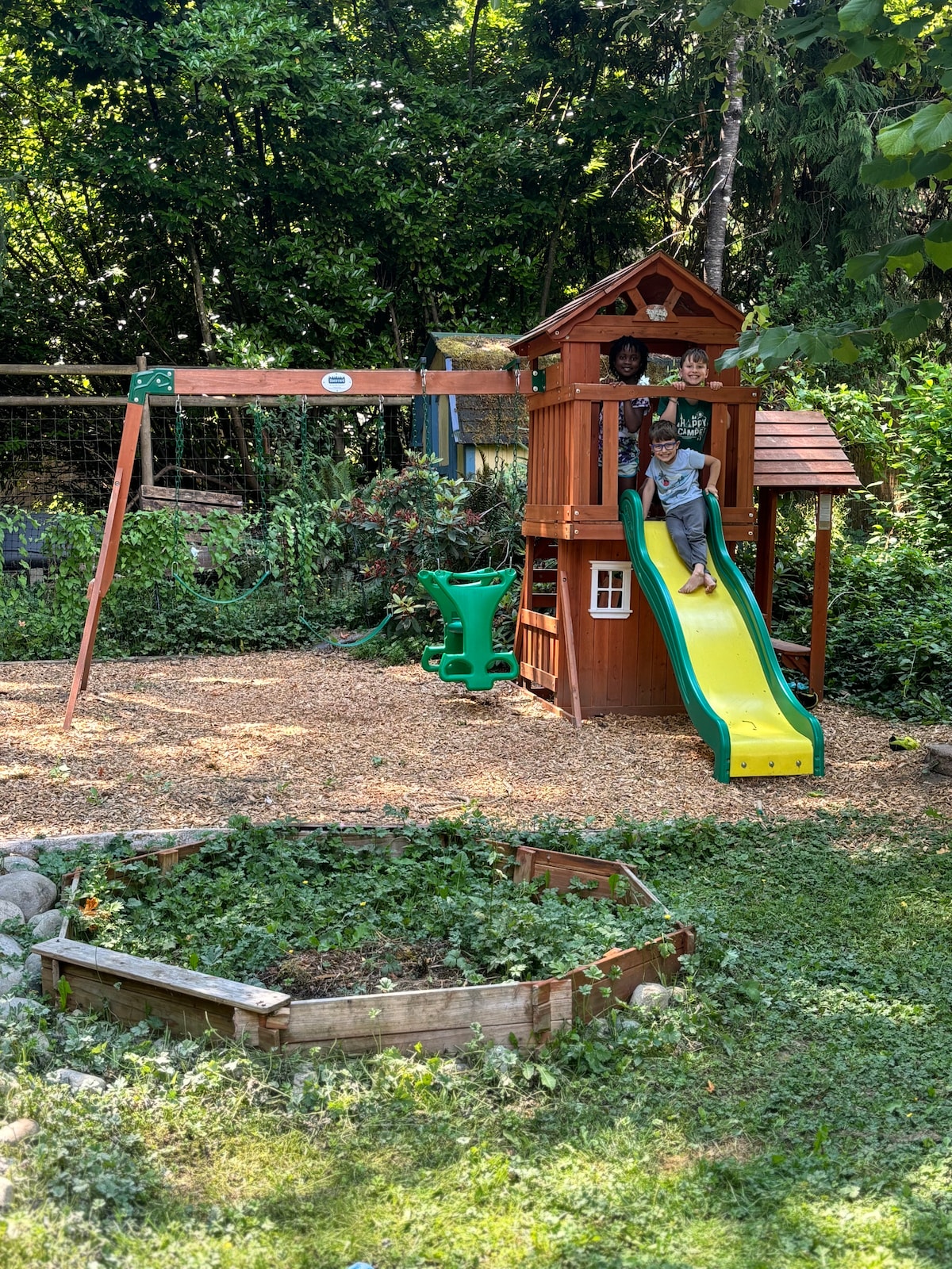 An outdoor play area features a wooden playset with a yellow slide and swings. The ground is covered in wood chips, and lush greenery surrounds the space. A circular garden bed made of wooden planks is visible in the foreground, filled with ground cover plants.
