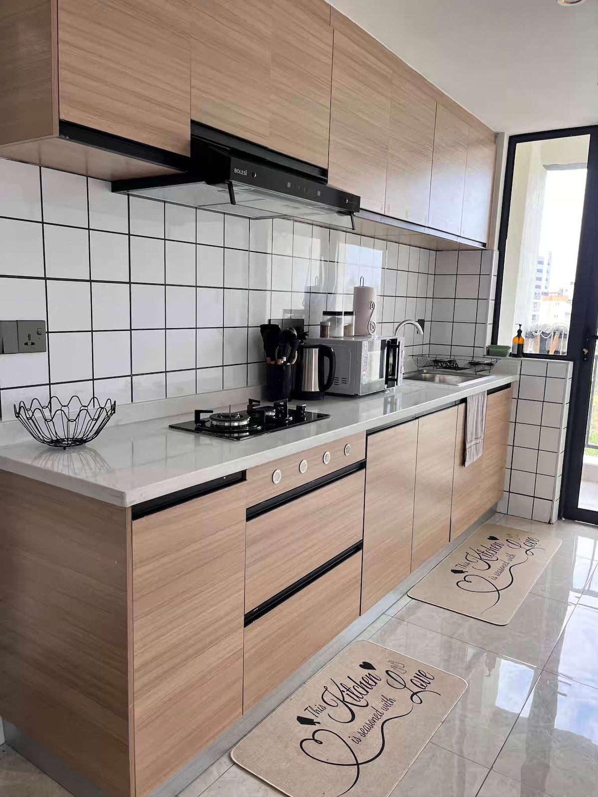 A modern kitchen features sleek cabinetry with a light wood finish, complemented by a white tiled backsplash. Cooking appliances include a gas stove and an oven. A coffee maker and kettle are visible on the countertop, while the floor showcases two decorative mats.