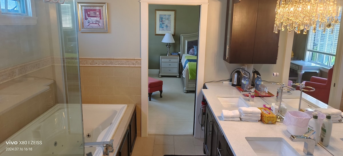 An inviting bathroom features a modern design with a glass-enclosed shower and a separate soaking tub. A double sink vanity is equipped with neatly arranged towels and personal care items. A doorway is visible, leading to a well-furnished bedroom, enhancing the suite's spacious layout.
