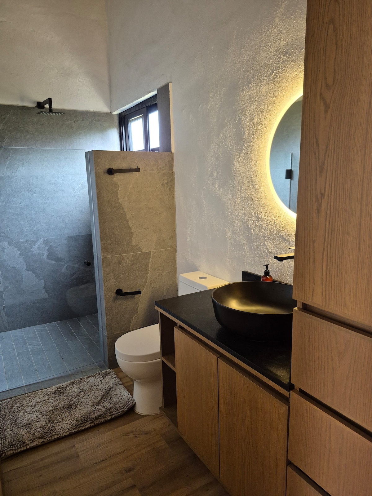 A modern bathroom is presented with a sleek shower featuring stone walls and a glass door. A bowl sink is positioned on a dark countertop. Natural light is allowed in through a window beside the mirror, and a soft rug is placed on the floor.