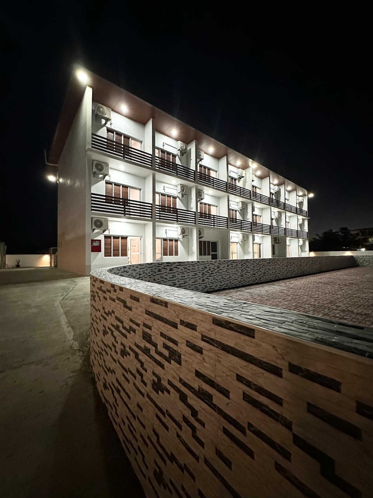 The exterior of the multilevel Proximity Apartments is showcased under night lighting, highlighting its modern architecture and design. Air conditioning units are visible on each level, while individual balconies add an inviting touch. A textured stone wall is seen in the foreground, complementing the structure.