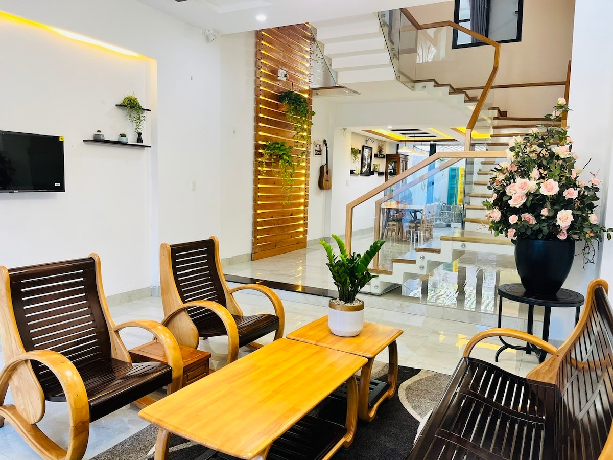 A spacious living area is featured with wooden chairs and a coffee table. A plant sits in the center of the table. Light wood accents and indoor greenery create a welcoming environment. Stairs in the background lead to the upper levels of the home.