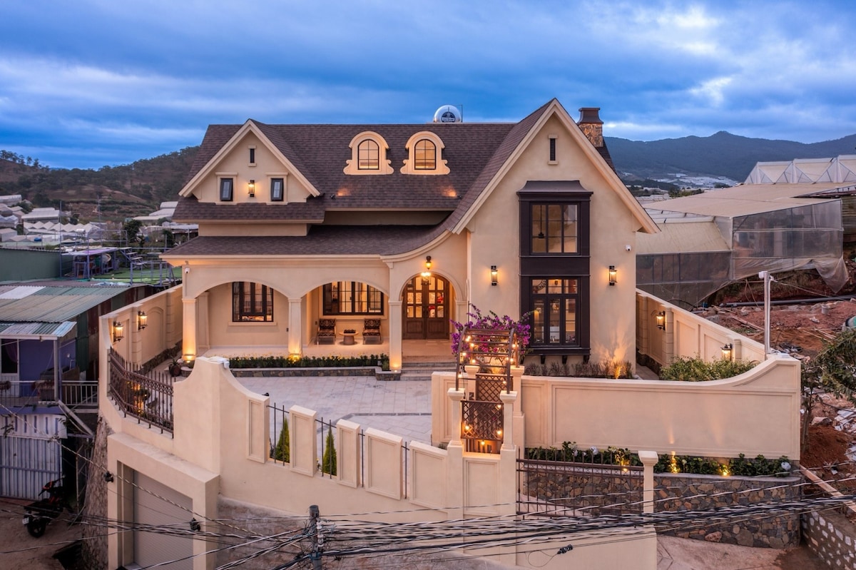 The villa's exterior showcases a charming architectural design with a combination of soft earth tones and elegant features. Warm lighting highlights the front entrance and outdoor patio area, which is surrounded by a low stone wall. A scenic mountain backdrop is visible in the distance.