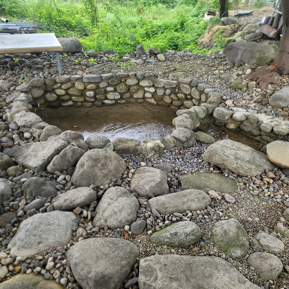 A natural stone hot tub is surrounded by a variety of smooth and textured rocks. The water reflects the greenery of the surrounding area, creating a serene outdoor setting. The scene is complemented by pebbles that form a pathway leading to the tub.