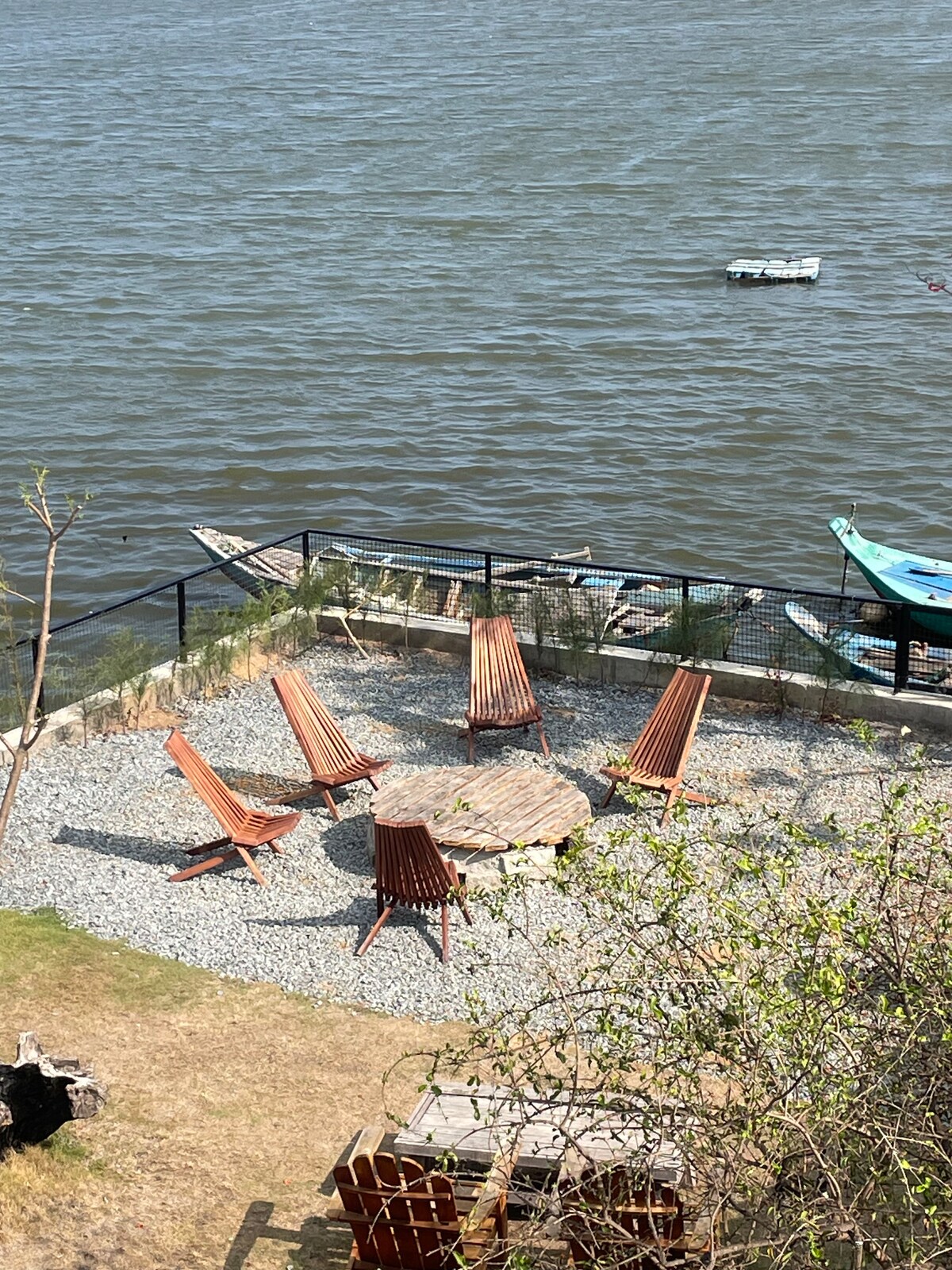 An outdoor seating area is surrounded by a gravel path, featuring six wooden chairs arranged around a circular table. The area overlooks a body of water, with boats visible in the distance and greenery along the edge.