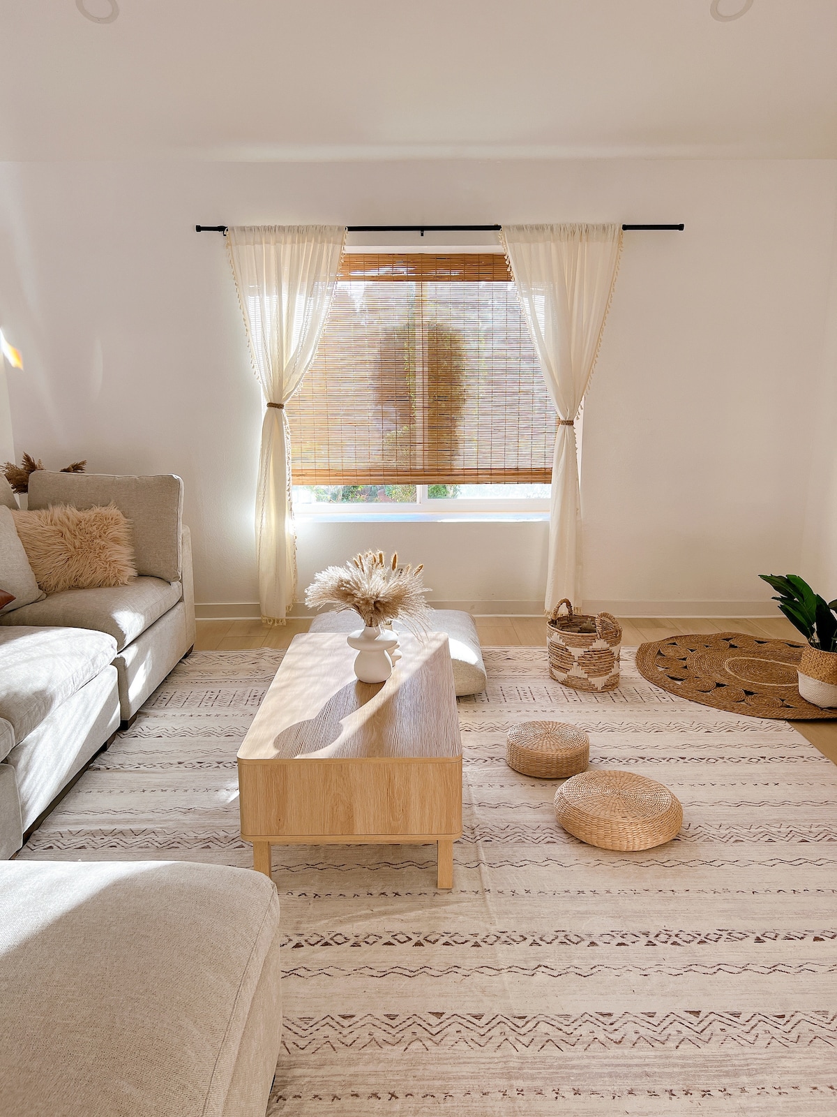 A bright living space is showcased with light filtering through bamboo shades. A neutral-colored sofa sits beside a wooden coffee table, adorned with a decorative vase. Textured poufs and a woven area rug add warmth, while plants introduce a touch of nature.