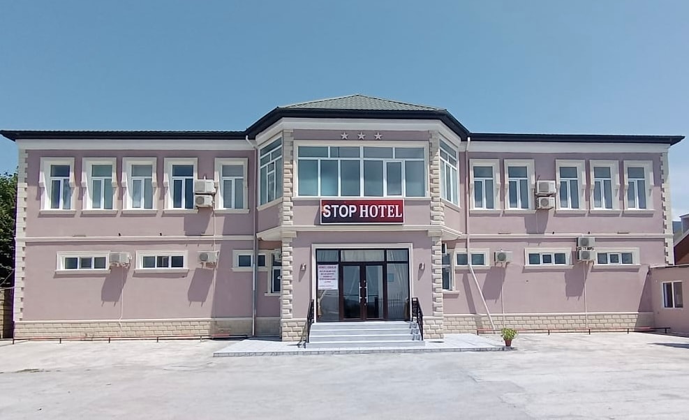 The exterior of the hotel features a light-colored façade with a modern design. Large glass windows provide natural light to the building, while a set of stairs leads to the entrance. Air conditioning units are visible on the walls, emphasizing comfort.