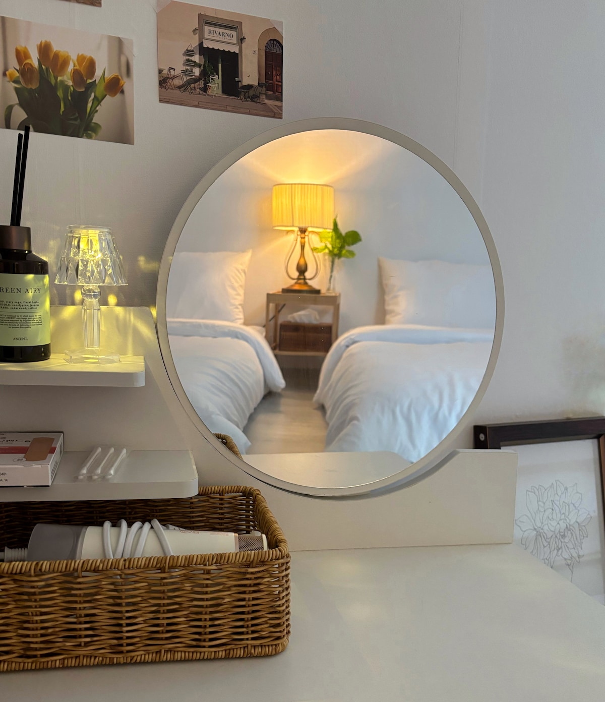 A round mirror reflects a bedside table featuring a lamp and a small plant. In the background, a spacious bed with white linens is visible, creating a calm and inviting space. Decorative items are neatly arranged on the shelf below the mirror.