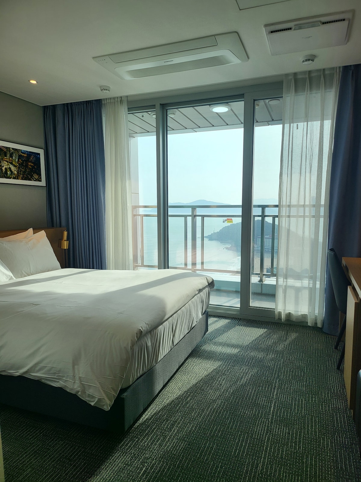 A bright room features a spacious bed covered with white linens. Floor-to-ceiling glass doors open to a balcony with views of distant hills and calm waters. Sheer curtains allow natural light to fill the space, complemented by a simple desk and chair.