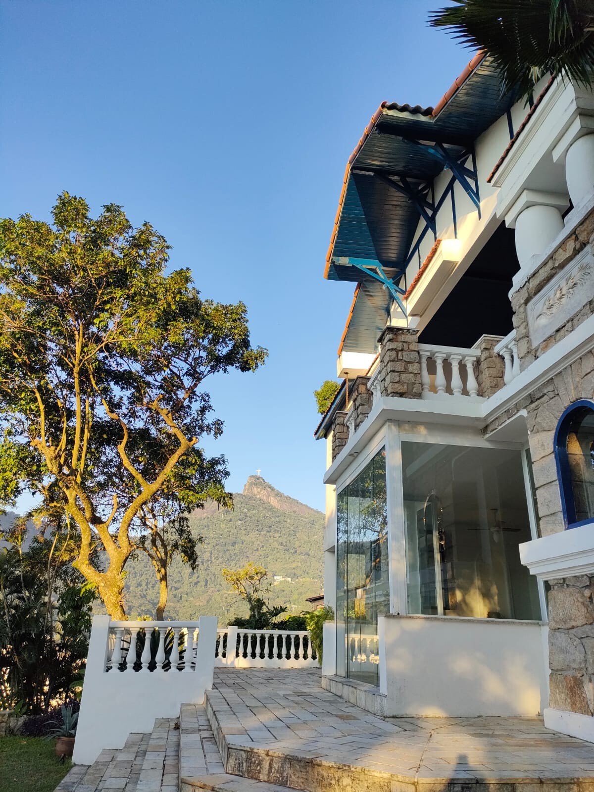 The exterior of the colonial-style house features a stone façade and large glass windows, surrounded by lush greenery. A staircase leads to an elevated patio area, providing views of the nearby mountainous landscape under clear blue skies.