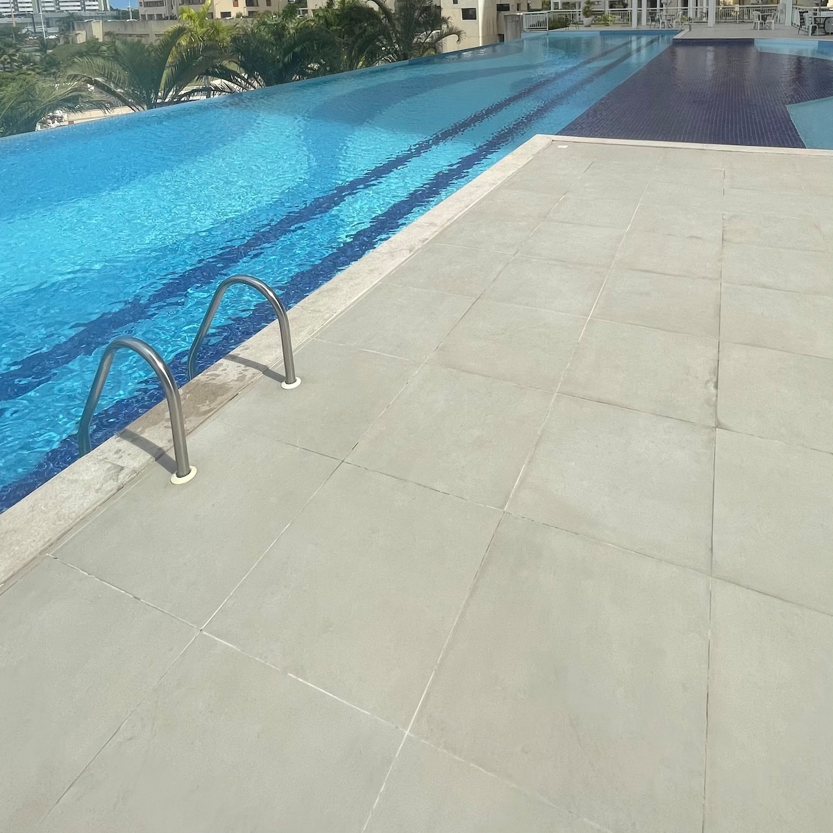 A lengthy swimming pool features calm, inviting waters, bordered by a smooth, grey tiled deck. Two stainless steel handrails are positioned for easy access. Lush greenery can be seen in the background, enhancing the serene ambiance of the area.