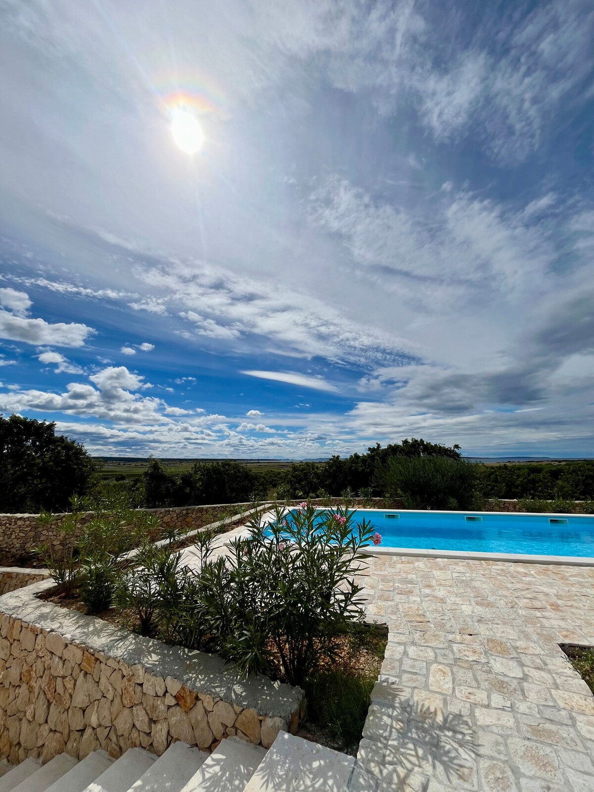 A serene outdoor space features a stone terrace with a swimming pool surrounded by greenery. The sky above is expansive, displaying clouds and sunlight. Scenic views of the olive grove and distant hills are visible in the background.