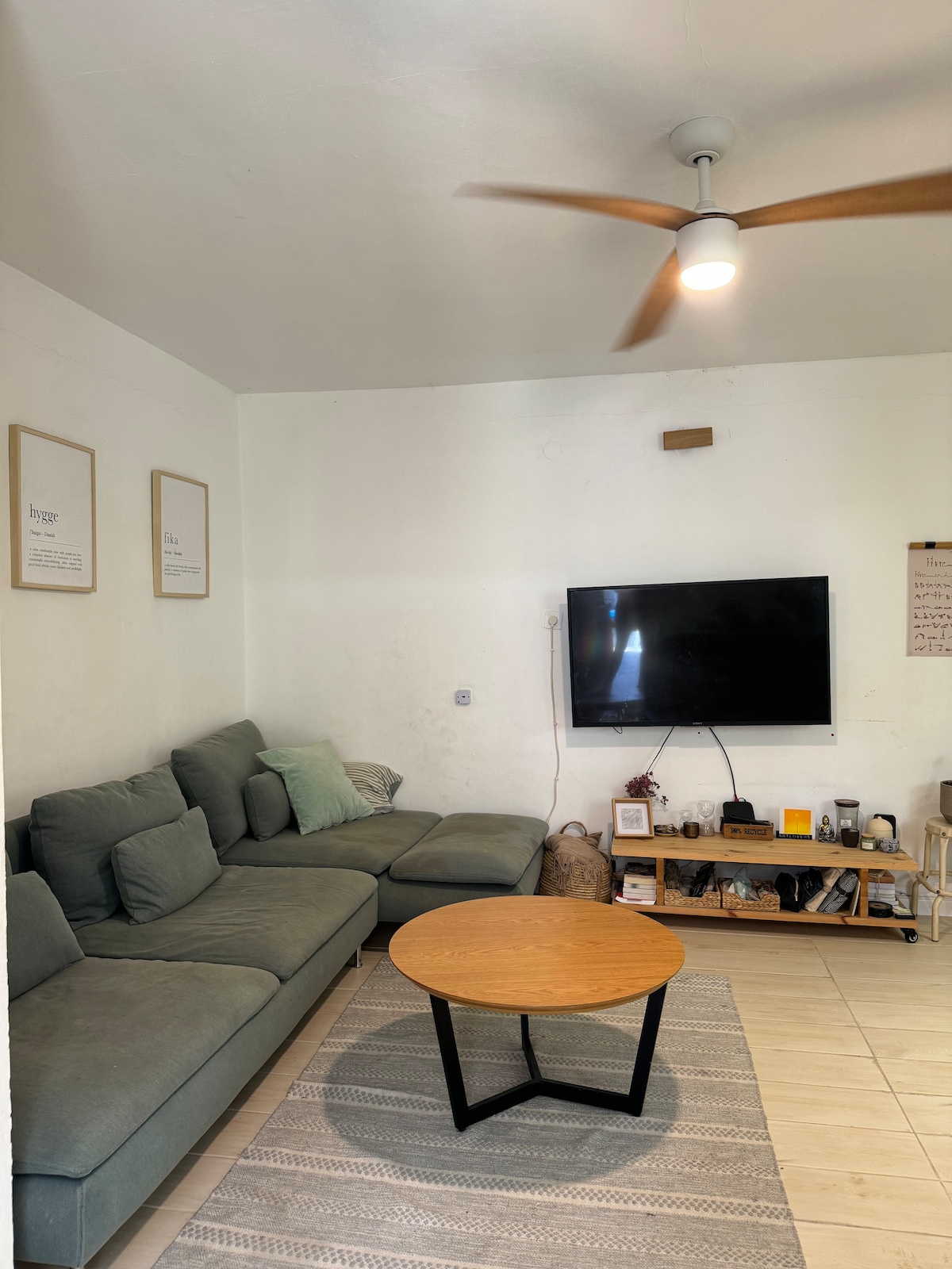 A modern living area features a large, L-shaped sofa in muted tones, complemented by a round wooden coffee table. A wall-mounted television is positioned across from the seating. Light-colored tiles contribute to the bright, airy feel of the space, while framed artwork adorns the walls.