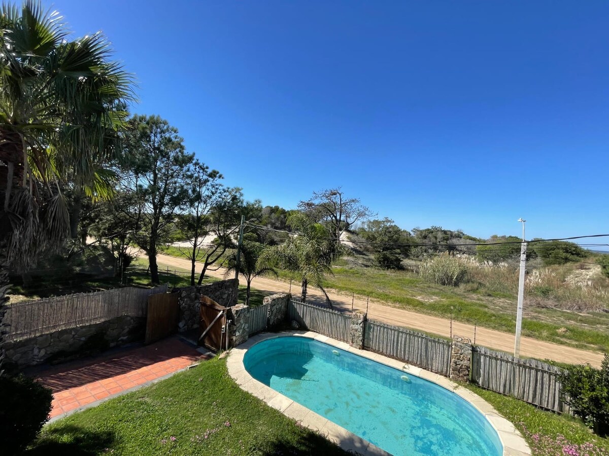 A view of the outdoor pool framed by lush greenery and a wooden fence. The clear blue sky is visible overhead, complementing the inviting water. Beyond the pool, a sandy road and natural landscape can be seen, enhancing the tranquil setting.