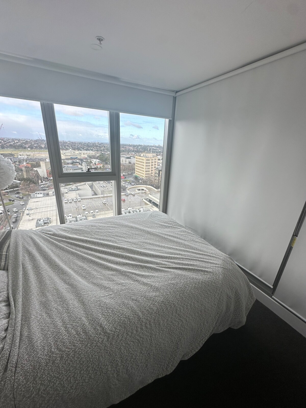 A bedroom offers a spacious bed draped in a light-colored coverlet, positioned against a large window that provides expansive views of the city skyline. Soft natural light enters, highlighting the clean, modern design of the room with neutral walls.