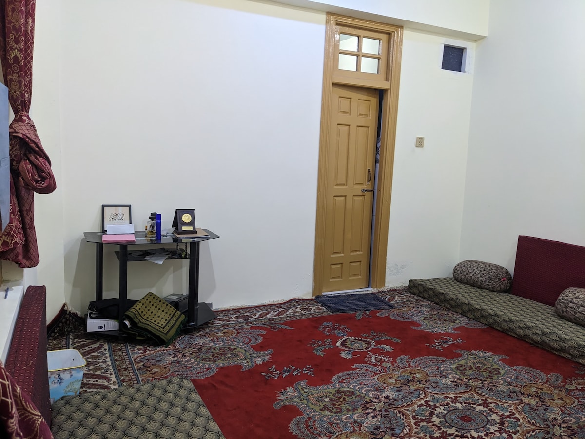 A cozy living area features a richly patterned red carpet with decorative designs. A small table holds various items, including a notebook and a trophy. The space includes low seating cushions along two walls, with a wooden door leading outside, allowing natural light to enter.