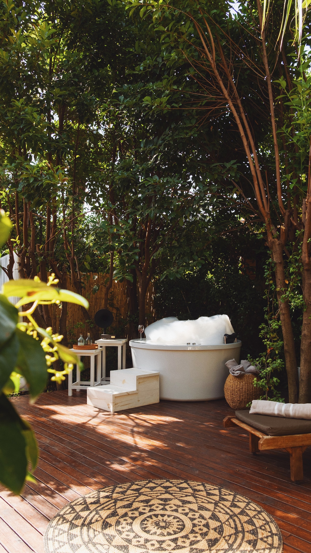 A serene outdoor space is enclosed by lush greenery, featuring a round white hot tub filled with bubbles. A small wooden table is positioned nearby, accompanied by a woven basket and a lounge chair on a wooden deck. A decorative rug enhances the tranquil setting.