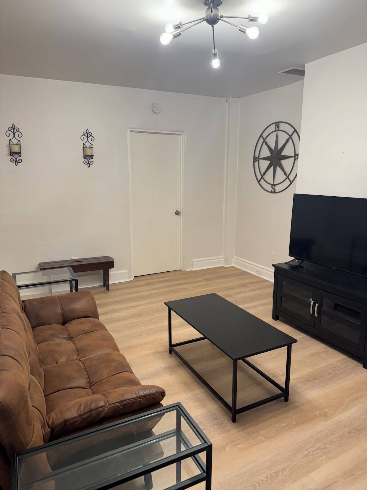 The living room features a brown futon adjacent to a black coffee table and a media console with a large flat-screen TV. A minimalist wall clock is displayed on one wall, while two decorative wall hangings occupy the opposite side. Natural light enhances the warm wood flooring.