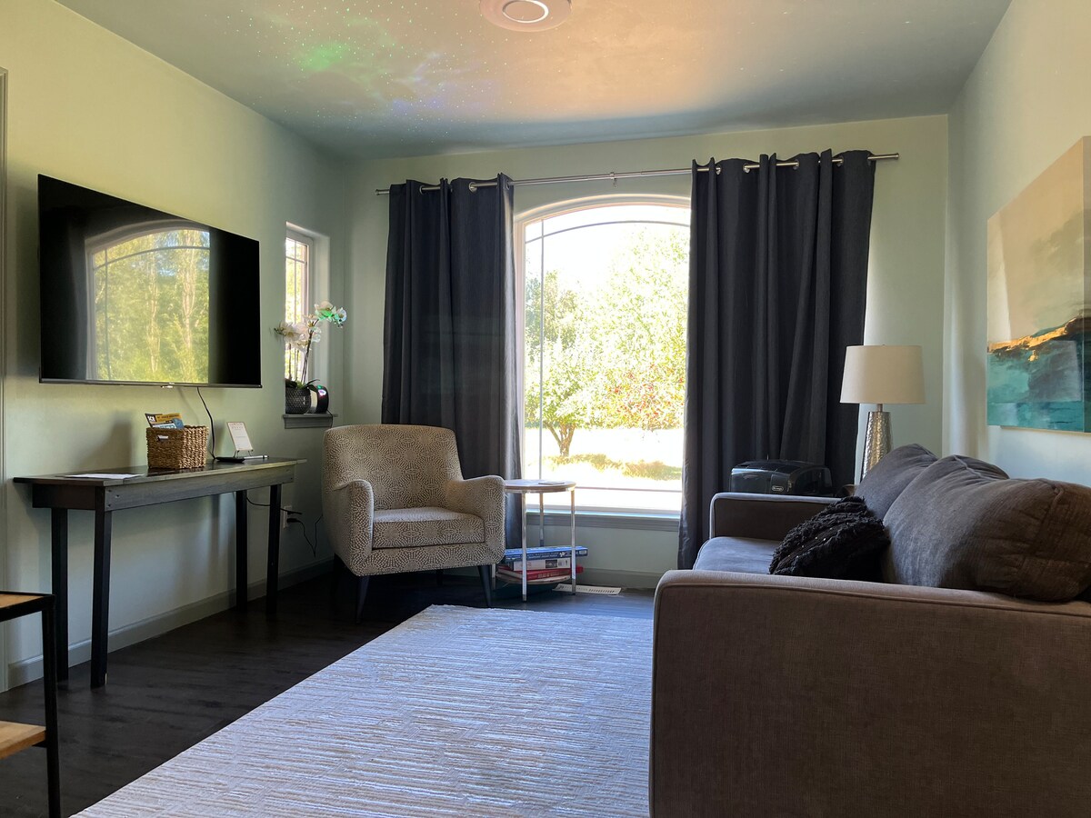 A living area is furnished with a comfortable sofa and an armchair positioned near a window. A media console with a television is mounted on the wall, and light filters through flowing curtains. An art piece is displayed on one wall, adding a touch of color.