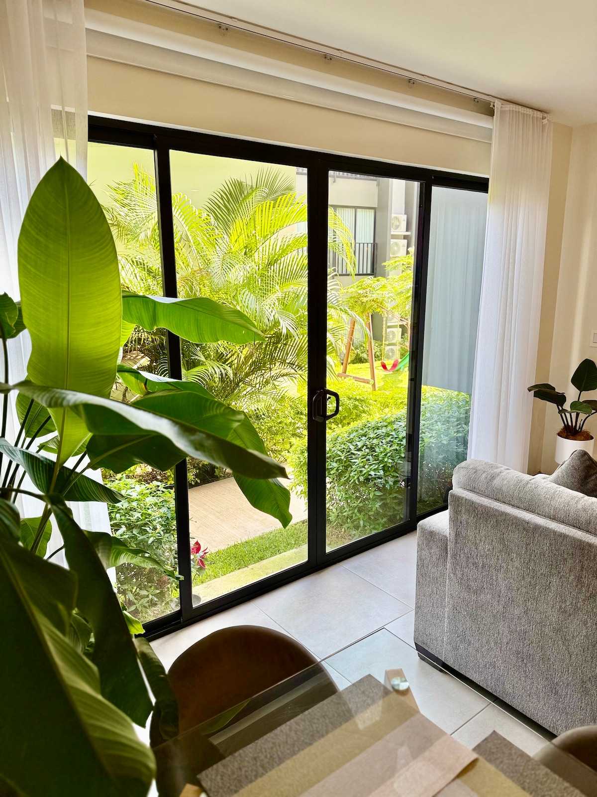 Large glass doors frame a view of a lush garden, featuring vibrant greenery and well-kept landscaping. Soft natural light filters through sheer curtains, creating a bright and inviting atmosphere in the room, complemented by indoor plants and a cozy gray sofa.