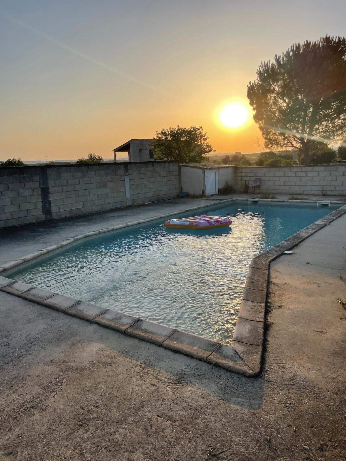 A swimming pool is featured, surrounded by a concrete deck. In the center, a colorful inflatable floats gently on the water's surface, reflecting the warm glow of the setting sun. The pool area is enclosed by a low stone wall, with trees visible beyond.