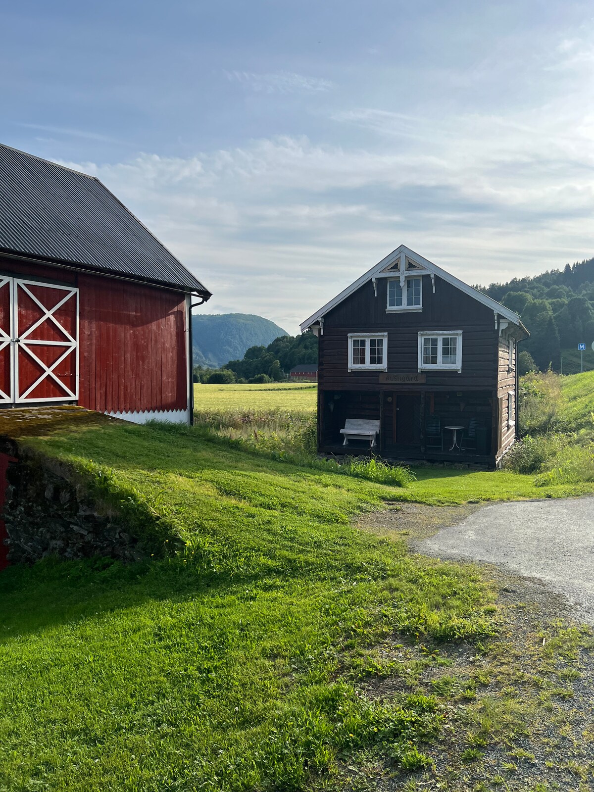 A two-story black house is positioned beside a red barn, surrounded by lush green grass. The landscape features gentle rolling hills in the background, with mountains visible under a clear blue sky. The area offers a peaceful rural setting.