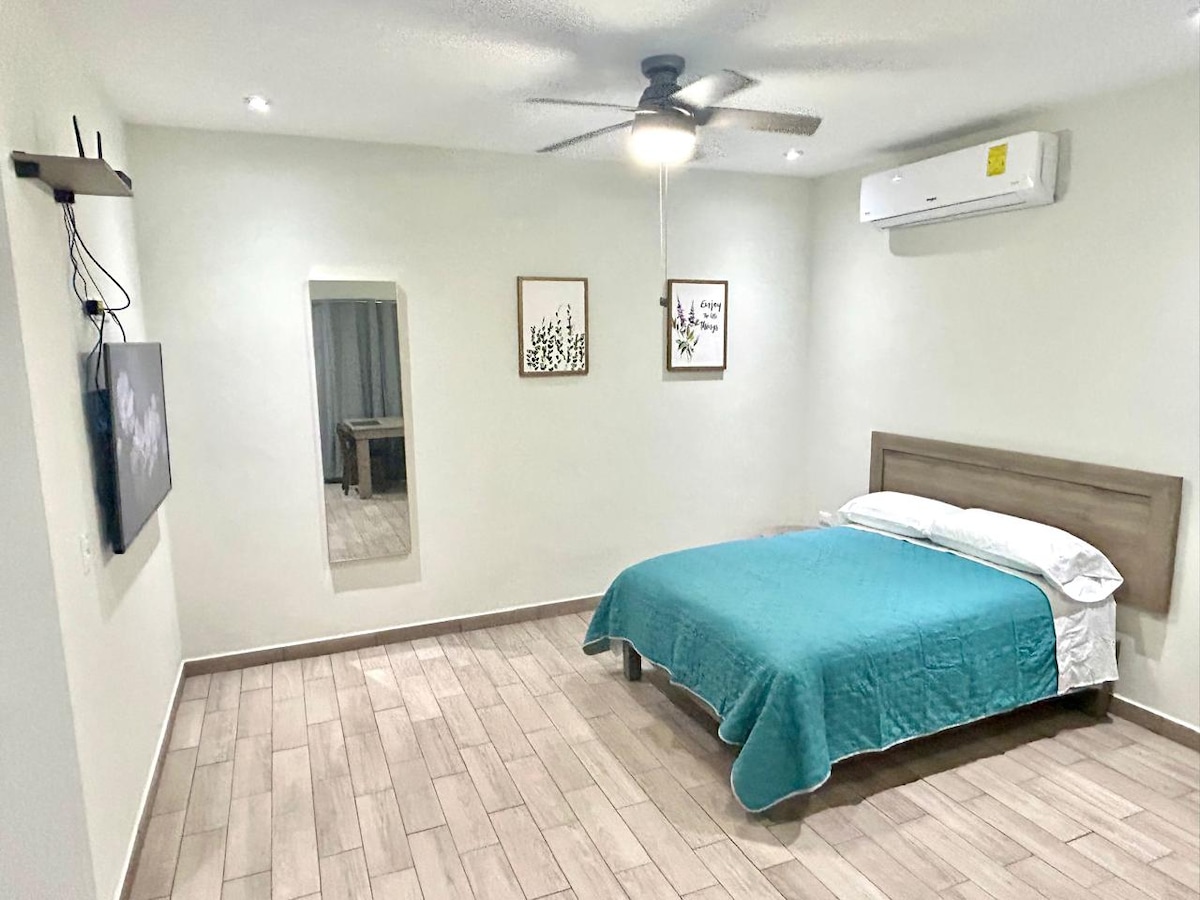 A well-lit bedroom features a spacious bed covered with a turquoise blanket. A ceiling fan is mounted above, while a television is mounted on the wall. A mirror and two framed artworks are displayed, adding to the minimalist decor.