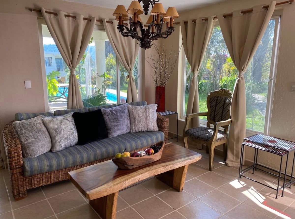 A living area is presented with a woven sofa adorned with various cushions, complemented by a wooden coffee table. Natural light enters through large windows, highlighting the serene outdoor view. A decorative chair and light fixtures add a touch of style to the space.