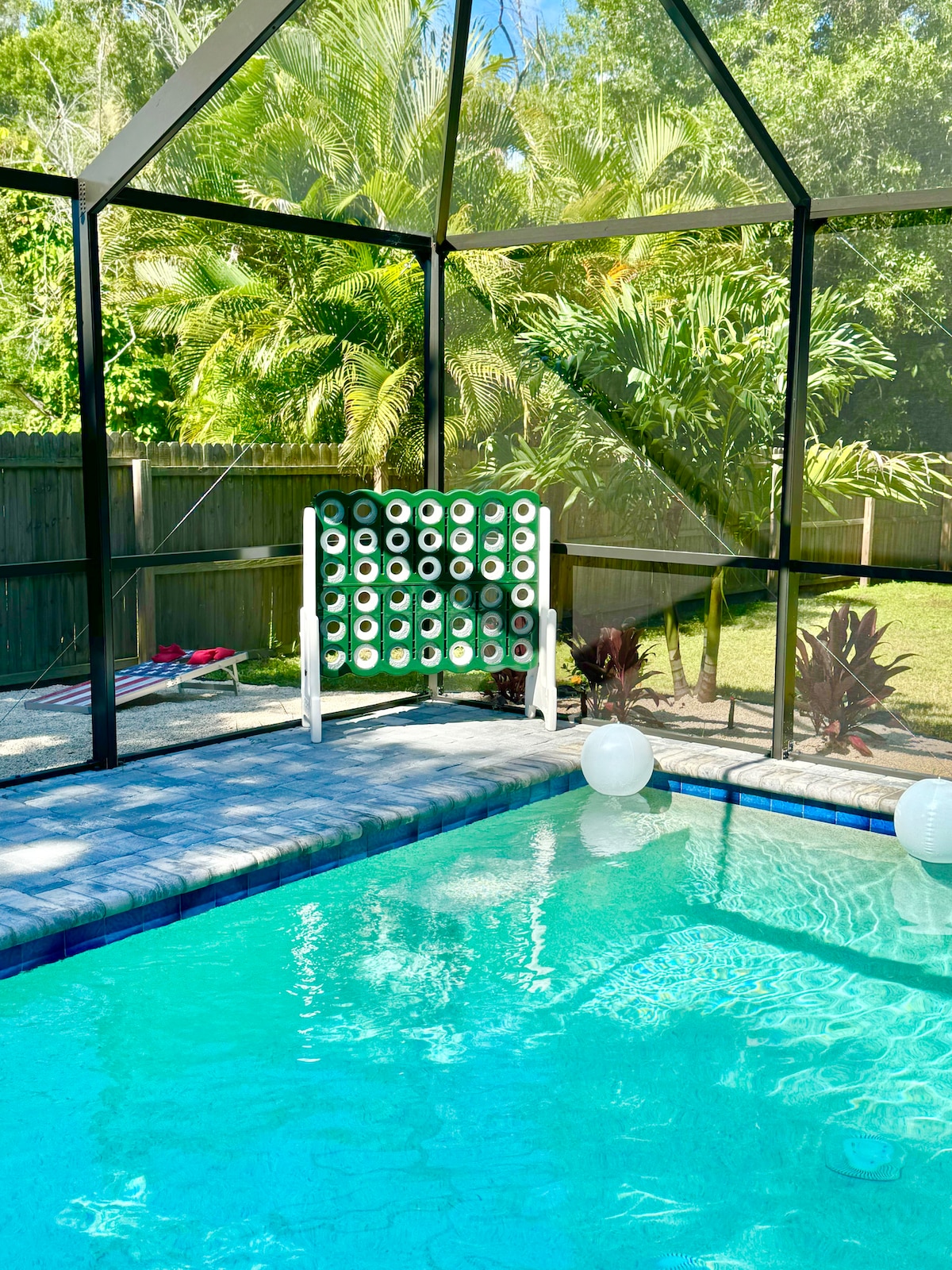 The screened-in area features a bright, heated saltwater pool, surrounded by lush tropical greenery. A large green game board is positioned nearby, with playful lighted orbs adding a subtle ambiance. The pool's clear water reflects the vibrant colors from the surrounding landscape.