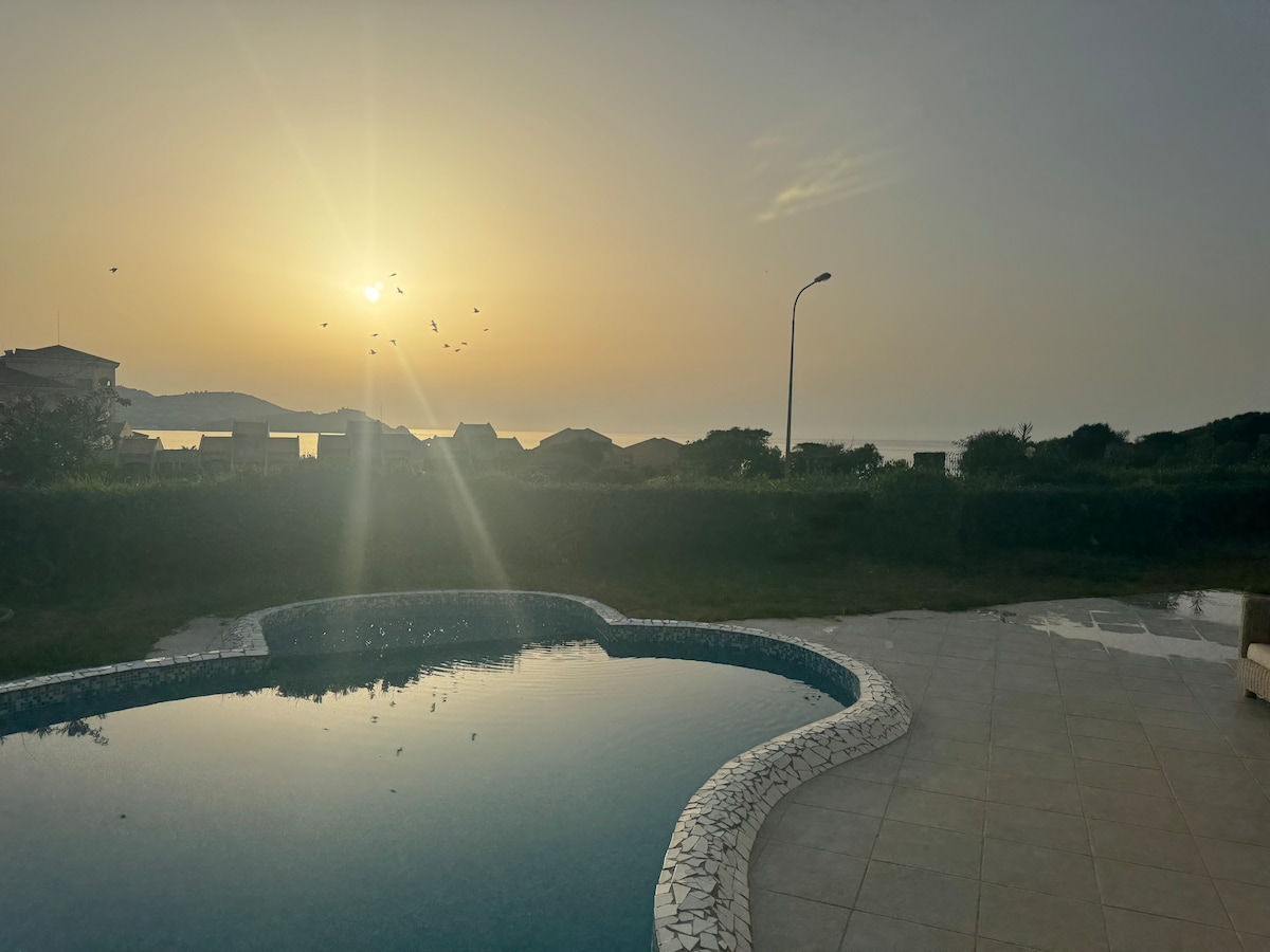 A tranquil outdoor space is highlighted by a serene pool reflecting the sunrise. Soft light creates a calming atmosphere, while distant silhouettes of houses and greenery are visible in the background. The area is surrounded by well-maintained landscaping, offering a peaceful retreat.