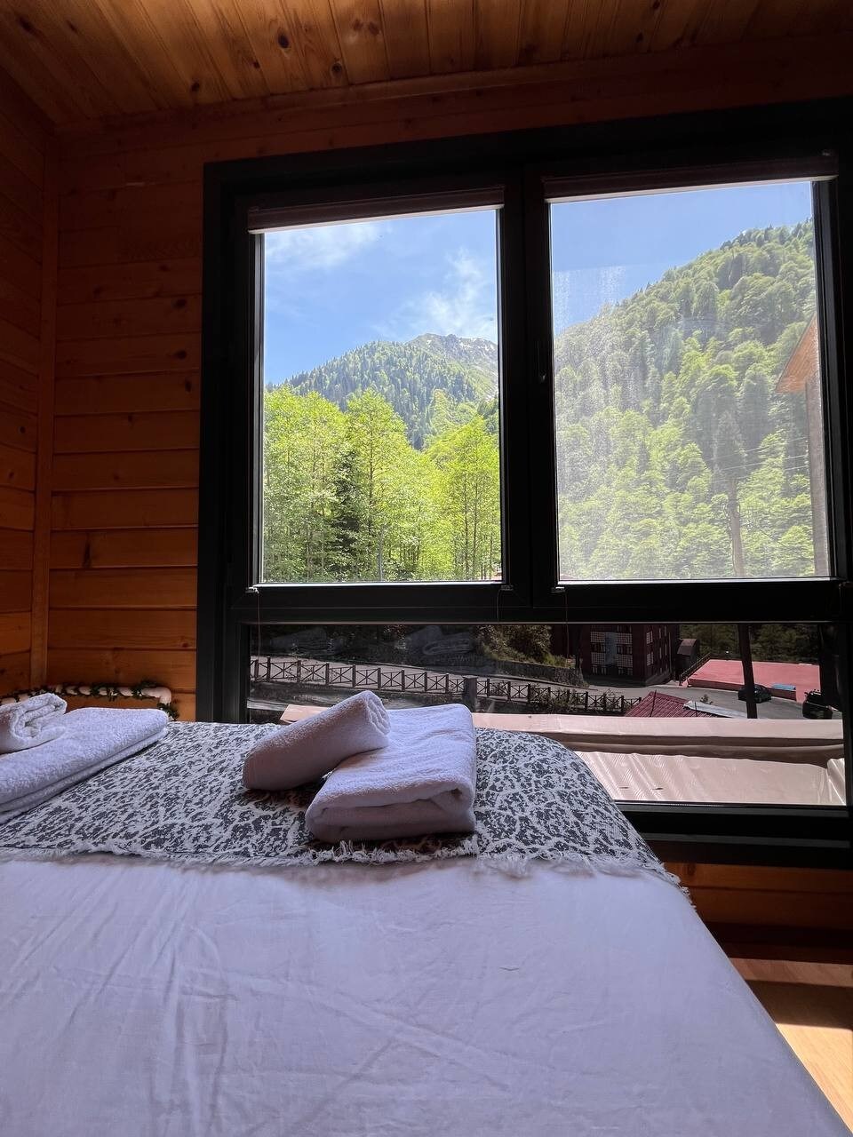 A cozy sleeping area features a spacious bed with a white blanket, accented by rolled towels. Large windows provide expansive views of lush green mountains and clear blue skies, filling the room with natural light.
