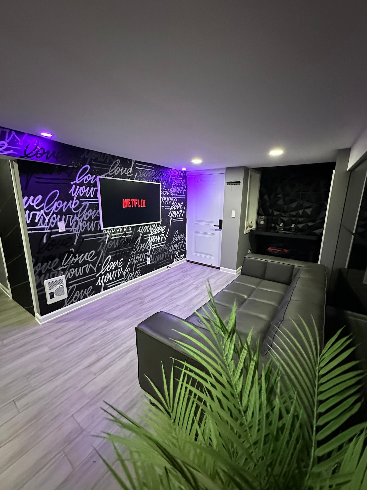A stylish living area is equipped with a sleek gray sofa, illuminated by ambient purple lighting. A wall adorned with text art displays the Netflix logo, enhancing the modern aesthetic. A large plant adds a touch of green, contributing to the inviting atmosphere.