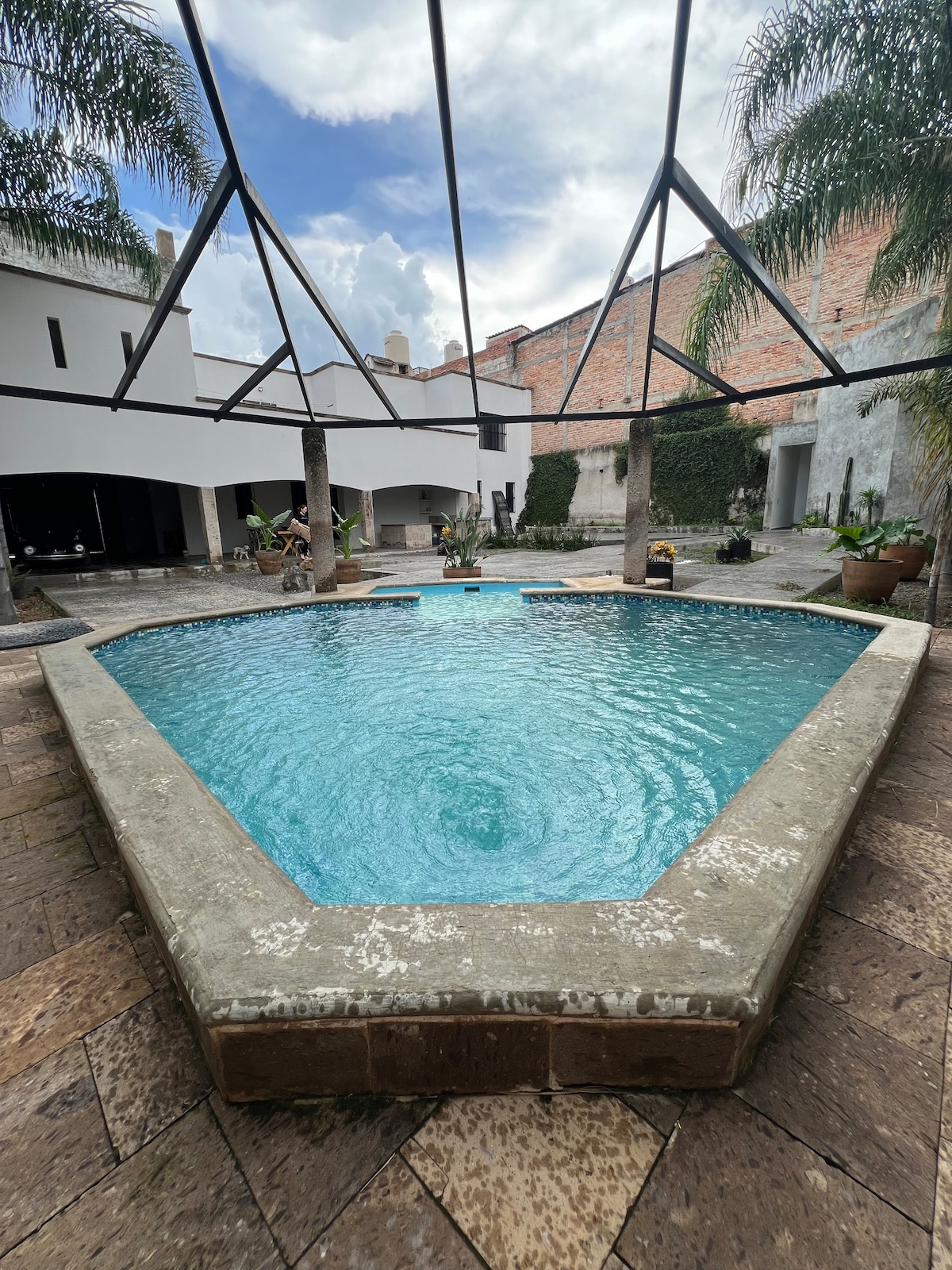 A triangular pool is surrounded by a stone deck, offering a refreshing space for relaxation. Palm trees are positioned at each corner, enhancing the tranquil atmosphere. The area is partially covered by a geometric metal structure, allowing natural light to filter in while providing shade.