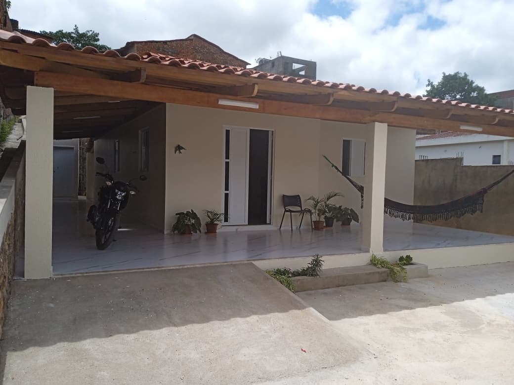 A covered front porch is highlighted by a hanging hammock and potted plants. A single chair is positioned near the entrance. A motorcycle is parked to the side, while the exterior features a light color scheme and modern windows that invite natural light.