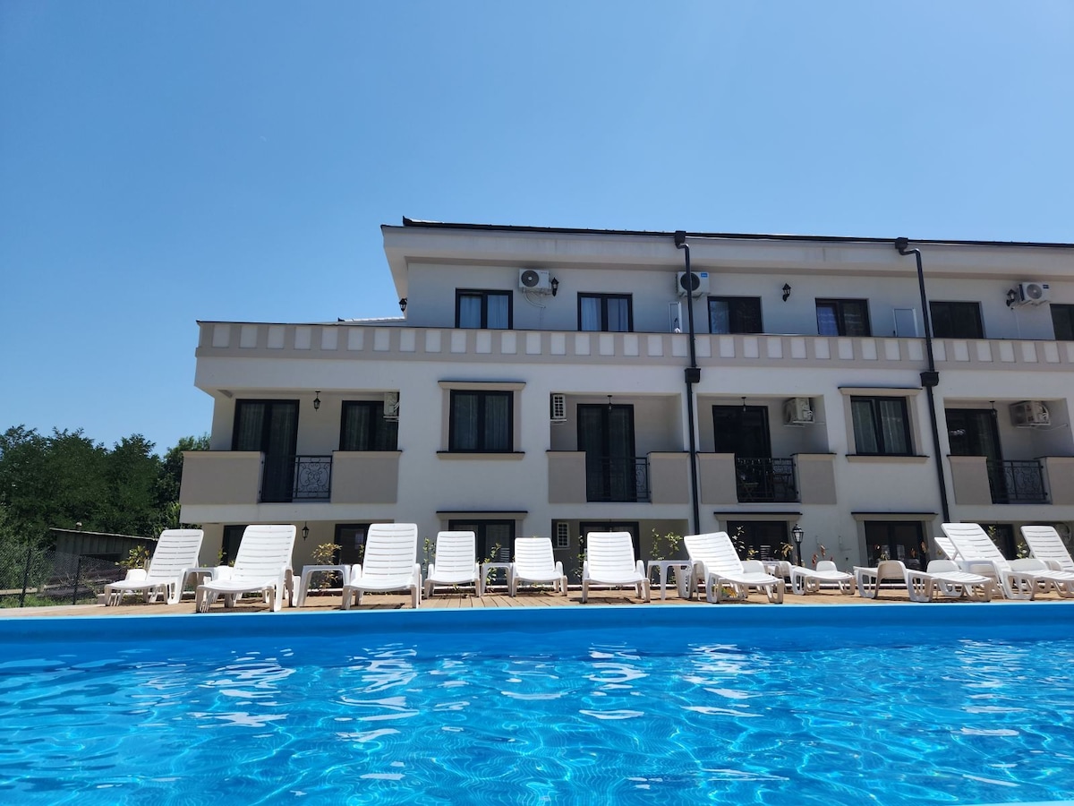 The image displays a modern building with multiple balconies and large windows, situated beside a bright blue swimming pool. White lounge chairs are positioned around the pool, creating a welcoming space for relaxation. Clear skies and ample sunlight enhance the serene atmosphere.