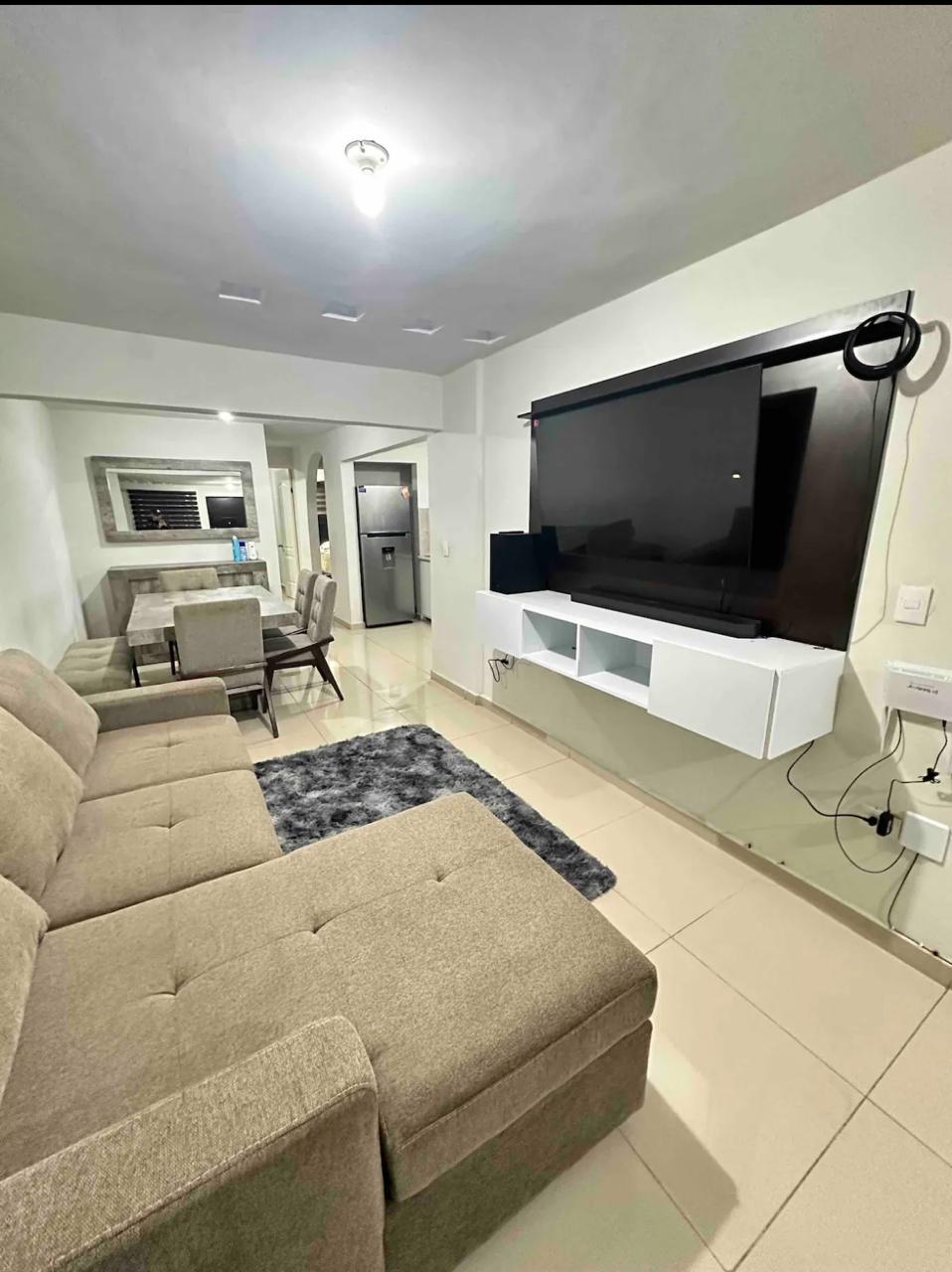 A modern living area features a comfortable sectional sofa facing a large wall-mounted television. A plush gray rug lies beneath the furniture. In the background, a dining table is visible, set for four, with a kitchen area beyond it.
