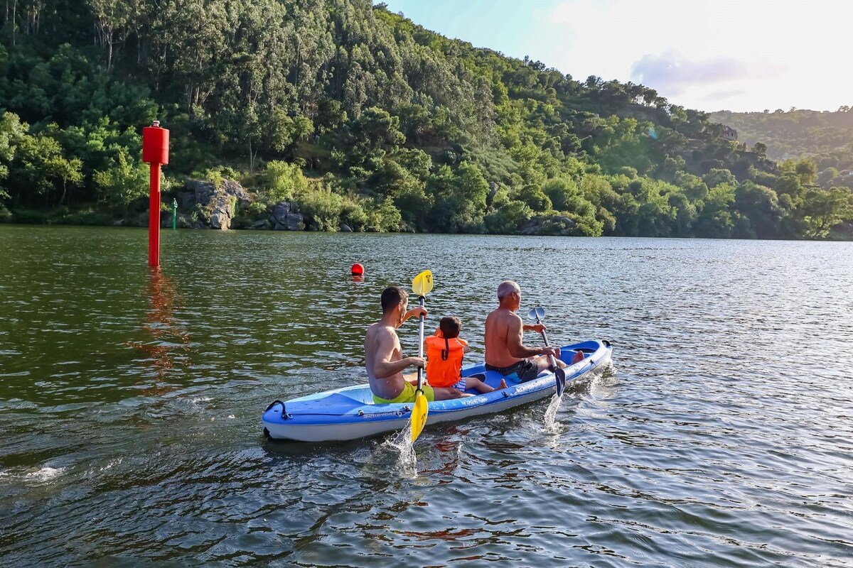 A kayak with three individuals glides smoothly across serene waters, surrounded by lush greenery. The sun reflects off the water's surface, and nearby, red navigation buoys mark the area. The scene captures a peaceful moment of outdoor recreation in a natural setting.