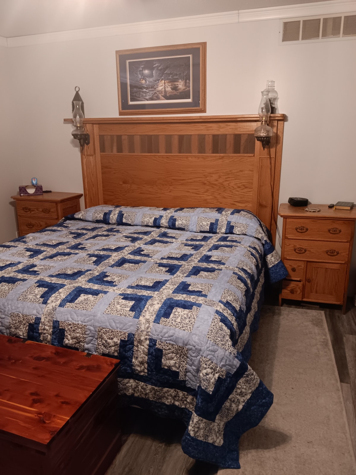 A wooden headboard frames a spacious bed adorned with a blue and white quilt. Matching bedside tables are positioned on either side, complemented by wall sconces. A piece of artwork hangs above the bed, adding a touch of character to the room.